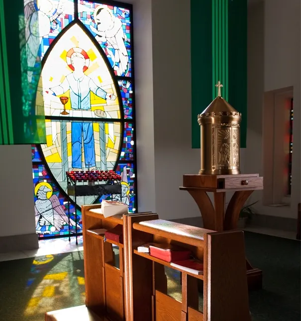 Sunlight shines through stained glass casting colorful light on kneelers and a tabernacle.